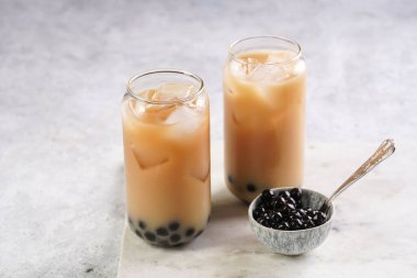 Two glasses with black tea, milk, ice cubes and cooked tapioca pearls for trendy bubble boba ice tea, small grey ceramic bowl on marble board on pastel tropical background