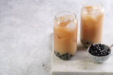 Two glasses with black tea, milk, ice cubes and cooked tapioca pearls for trendy bubble boba ice tea, two small grey ceramic bowls on marble board on pastel grey background