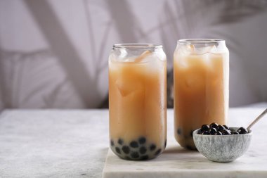 Two glasses with black tea, milk, ice cubes and cooked tapioca pearls for trendy bubble boba ice tea, two small grey ceramic bowls on marble board on pastel tropical background, cat in the back
