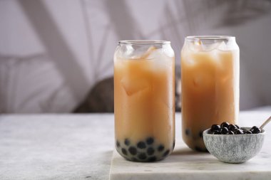 Two glasses with black tea, milk, ice cubes and cooked tapioca pearls for trendy bubble boba ice tea, two small grey ceramic bowls on marble board on pastel tropical background, cat in the back