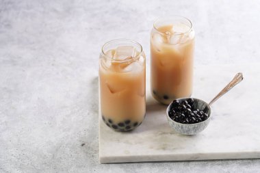Two glasses with black tea, milk, ice cubes and cooked tapioca pearls for trendy bubble boba ice tea, two small grey ceramic bowls on marble board on pastel grey background