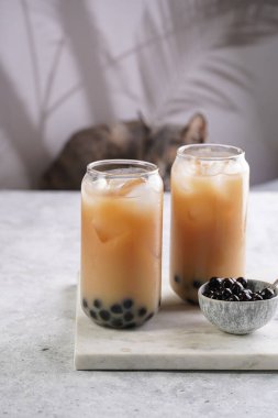 Two glasses with black tea, milk, ice cubes and cooked tapioca pearls for trendy bubble boba ice tea, two small grey ceramic bowls on marble board on pastel tropical background, cat in the back