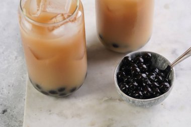 Two glasses with black tea, milk, ice cubes and cooked tapioca pearls for trendy bubble boba ice tea, two small grey ceramic bowls on marble board on pastel grey background