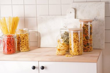 Variety of dry italian pasta in glass jars on kitchen cabinet with wooden top: fusilli, tagliatelle, farfalle, spaghetti