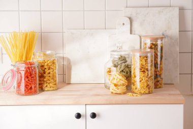 Variety of dry italian pasta in glass jars on kitchen cabinet with wooden top: fusilli, tagliatelle, farfalle, spaghetti