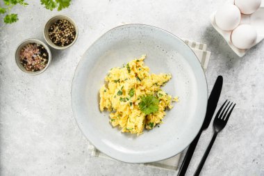 Kahvaltıda çırpılmış yumurta, yeşil otlar, gri masalarda maydanoz, siyah çatal ve bıçak, üst görüş.