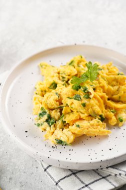 Kahvaltıda çırpılmış yumurta, yeşil otlar, beyaz tabakta maydanoz, gri masa, altın çatal ve bıçak.