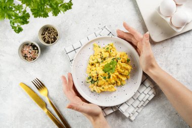 Kahvaltıda çırpılmış yumurta ve yeşil otlar tutan kadın eller, gri masadaki maydanoz, altın çatal ve bıçak, üst görüş.