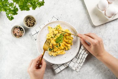 Kahvaltıda çırpılmış yumurta ve yeşil otlar tutan kadın eller, gri masadaki maydanoz, altın çatal ve bıçak, üst görüş.