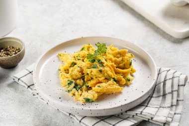 Kahvaltıda çırpılmış yumurta, yeşil otlar, beyaz tabakta maydanoz, gri masa, altın çatal ve bıçak.