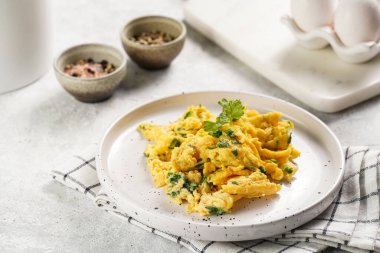 Kahvaltıda çırpılmış yumurta, yeşil otlar, beyaz tabakta maydanoz, gri masa, altın çatal ve bıçak.