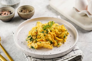 Kahvaltıda çırpılmış yumurta, yeşil otlar, beyaz tabakta maydanoz, gri masa, altın çatal ve bıçak.