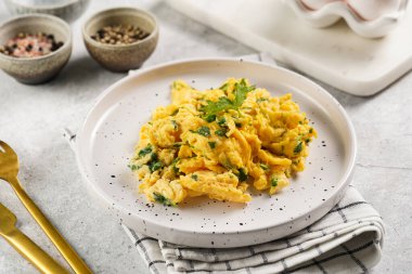 Kahvaltıda çırpılmış yumurta, yeşil otlar, beyaz tabakta maydanoz, gri masa, altın çatal ve bıçak.