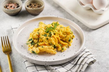 Kahvaltıda çırpılmış yumurta, yeşil otlar, beyaz tabakta maydanoz, gri masa, altın çatal ve bıçak.