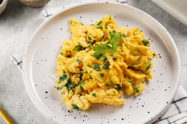 Kahvaltıda çırpılmış yumurta, yeşil otlar, beyaz tabakta maydanoz, gri masa, altın çatal ve bıçak.