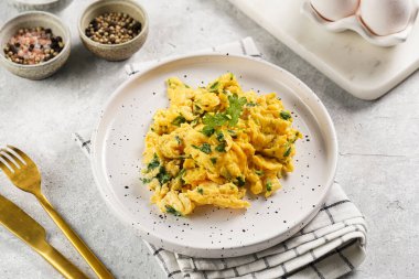 Kahvaltıda çırpılmış yumurta, yeşil otlar, beyaz tabakta maydanoz, gri masa, altın çatal ve bıçak.