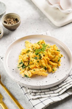 Kahvaltıda çırpılmış yumurta, yeşil otlar, beyaz tabakta maydanoz, gri masa, altın çatal ve bıçak.