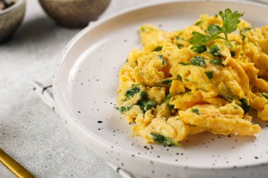 Kahvaltıda çırpılmış yumurta, yeşil otlar, beyaz tabakta maydanoz, gri masa, altın çatal ve bıçak.