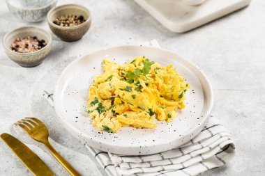 Kahvaltıda çırpılmış yumurta, yeşil otlar, beyaz tabakta maydanoz, gri masa, altın çatal ve bıçak.