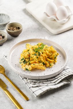 Kahvaltıda çırpılmış yumurta, yeşil otlar, beyaz tabakta maydanoz, gri masa, altın çatal ve bıçak.