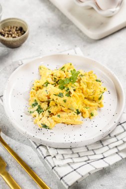 Kahvaltıda çırpılmış yumurta, yeşil otlar, beyaz tabakta maydanoz, gri masa, altın çatal ve bıçak.