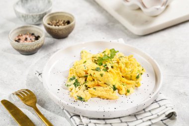 Kahvaltıda çırpılmış yumurta, yeşil otlar, beyaz tabakta maydanoz, gri masa, altın çatal ve bıçak.