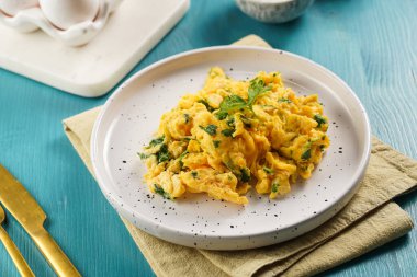 Kahvaltıda çırpılmış yumurta, yeşil otlar, maydanozlu beyaz tabak üzerinde canlı mavi ahşap masa, altın çatal ve bıçak.