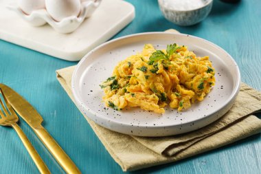 Kahvaltıda çırpılmış yumurta, yeşil otlar, maydanozlu beyaz tabak üzerinde canlı mavi ahşap masa, altın çatal ve bıçak.