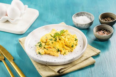 Kahvaltıda çırpılmış yumurta, yeşil otlar, maydanozlu beyaz tabak üzerinde canlı mavi ahşap masa, altın çatal ve bıçak.