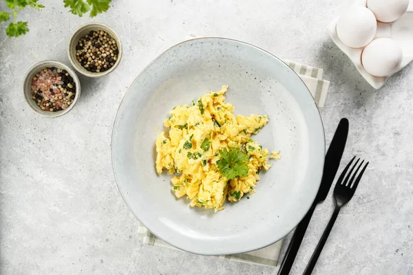 Kahvaltıda çırpılmış yumurta, yeşil otlar, gri masalarda maydanoz, siyah çatal ve bıçak, üst görüş.