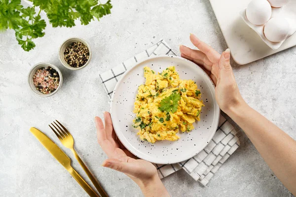 Kahvaltıda çırpılmış yumurta ve yeşil otlar tutan kadın eller, gri masadaki maydanoz, altın çatal ve bıçak, üst görüş.