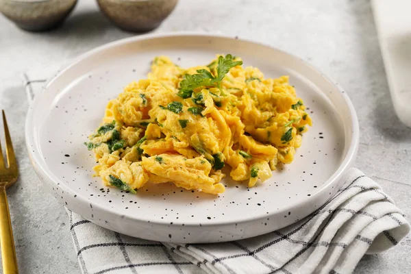 Kahvaltıda çırpılmış yumurta, yeşil otlar, beyaz tabakta maydanoz, gri masa, altın çatal ve bıçak.