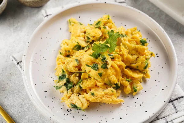 Kahvaltıda çırpılmış yumurta, yeşil otlar, beyaz tabakta maydanoz, gri masa, altın çatal ve bıçak.