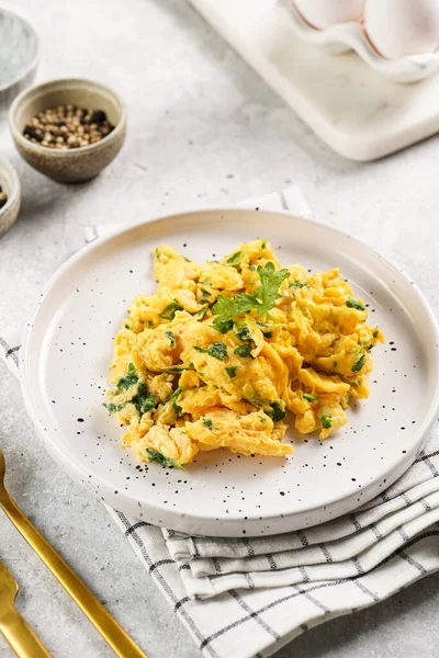 Kahvaltıda çırpılmış yumurta, yeşil otlar, beyaz tabakta maydanoz, gri masa, altın çatal ve bıçak.