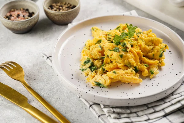 Kahvaltıda çırpılmış yumurta, yeşil otlar, beyaz tabakta maydanoz, gri masa, altın çatal ve bıçak.