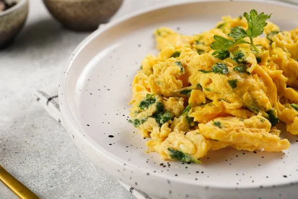 Kahvaltıda çırpılmış yumurta, yeşil otlar, beyaz tabakta maydanoz, gri masa, altın çatal ve bıçak.