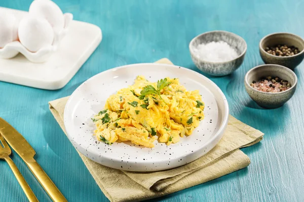 Kahvaltıda çırpılmış yumurta, yeşil otlar, maydanozlu beyaz tabak üzerinde canlı mavi ahşap masa, altın çatal ve bıçak.