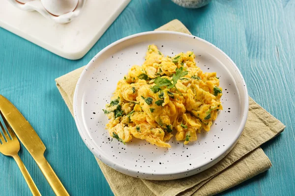 Kahvaltıda çırpılmış yumurta, yeşil otlar, maydanozlu beyaz tabak üzerinde canlı mavi ahşap masa, altın çatal ve bıçak.