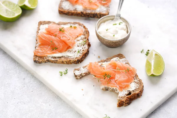 bitten off Wholewheat bread sandwiches with cream cheese and smoked salmon on marble board with lime slices and sour cream