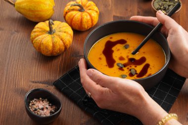 Elinde turuncu kabak kremalı bir kase ve kabak tohumu yağlı iki siyah kaşık tutan kadın elleri.