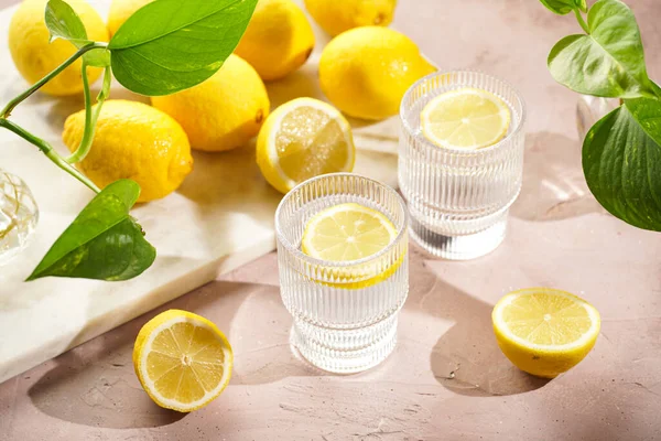 朝の飲み物 新鮮な柑橘類のスライスとレモン水の2つのガラス 淡いピンクの背景に多くのレモン全体 — ストック写真