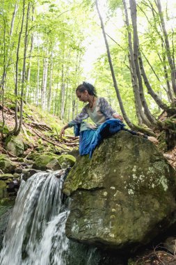 Mavi elbiseli esmer kadın bir dağ şelalesinin yanında oturuyor, Karpat yaz ormanı, Ukrain.