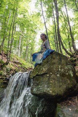Mavi elbiseli esmer kadın bir dağ şelalesinin yanında oturuyor, Karpat yaz ormanı, Ukrain.