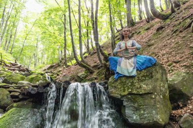 Mavi elbiseli esmer kadın bir dağ şelalesinin yanında oturuyor, Karpat yaz ormanı, Ukrain.