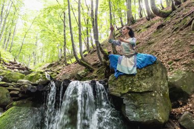Mavi elbiseli esmer kadın bir dağ şelalesinin yanında oturuyor, Karpat yaz ormanı, Ukrain.
