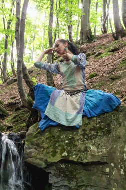 Mavi elbiseli esmer kadın bir dağ şelalesinin yanında oturuyor, Karpat yaz ormanı, Ukrain.