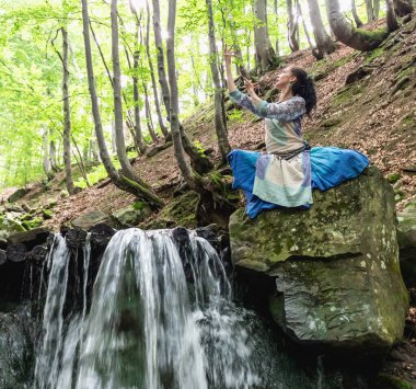 Mavi elbiseli esmer kadın bir dağ şelalesinin yanında oturuyor, Karpat yaz ormanı, Ukrain.