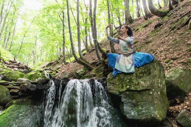 Mavi elbiseli esmer kadın bir dağ şelalesinin yanında oturuyor, Karpat yaz ormanı, Ukrain.