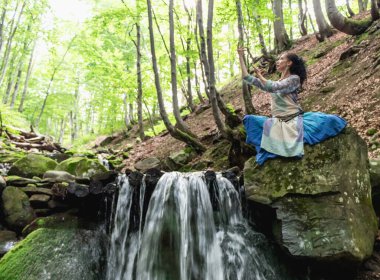 Mavi elbiseli esmer kadın bir dağ şelalesinin yanında oturuyor, Karpat yaz ormanı, Ukrain.