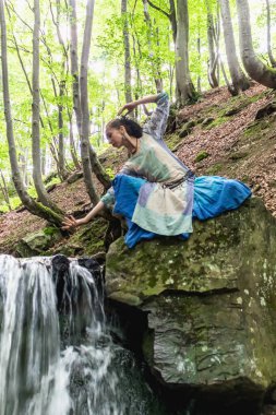 Mavi elbiseli esmer kadın bir dağ şelalesinin yanında oturuyor, Karpat yaz ormanı, Ukrain.